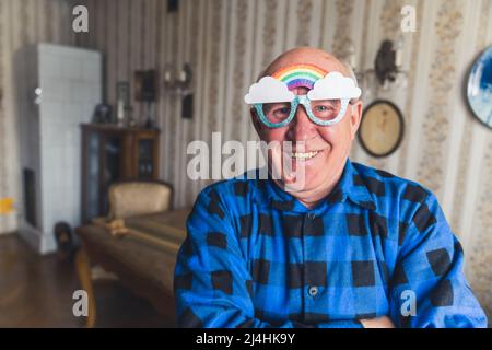 Un smiley funky caucasien ancien homme à la retraite dans une chemise en flanelle regardant la caméra, et souriant avec des lunettes arc-en-ciel. Soutien LGBT. Intérieur vintage. Photo de haute qualité Banque D'Images