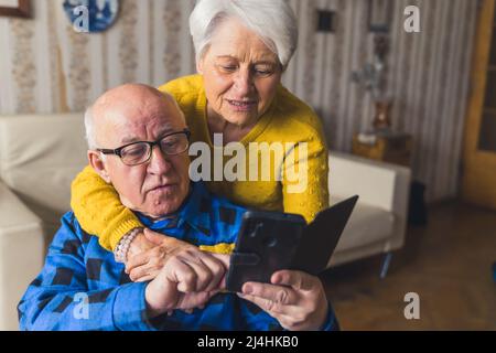 Femme caucasienne âgée à poil court embrassant son mari retraité depuis le dos et l'aidant avec son smartphone moderne. Intérieur de salon vintage. Photo de haute qualité Banque D'Images