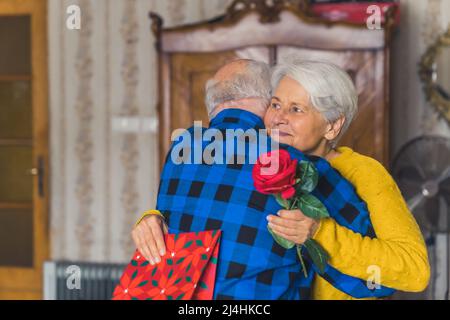 Concept de célébration romantique. Couple caucasien âgé célébrant leur anniversaire. Un homme à la retraite embrassant sa femme aux cheveux gris qui tient une rose rouge et un sac cadeau. Photo de haute qualité Banque D'Images