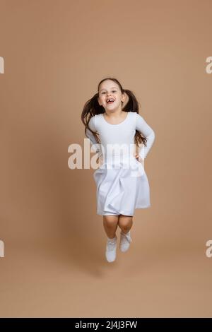 Portrait de jeune fille belle avec de longs cheveux foncés en robe blanche, chaussettes et chaussures de gymnastique saut, tenant les mains à la taille sur fond marron. Banque D'Images