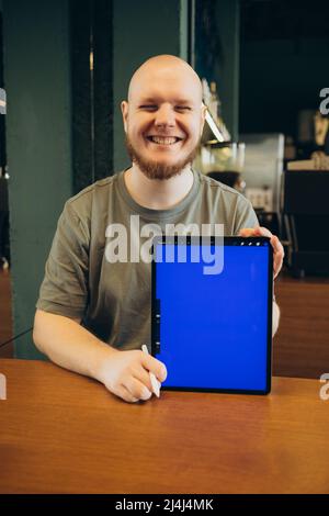 Un homme joyeux a pointé avec le doigt l'écran bleu vide de la tablette tout en étant assis sur le banc près d'un café moderne et en regardant la caméra Banque D'Images