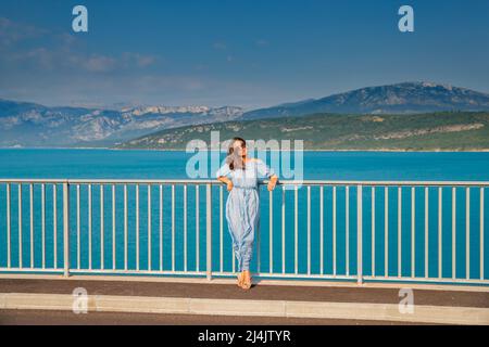 La belle fille dans une robe bleue et des lunettes de soleil pose sur le pont, les longs cheveux de châtaignier, heureux et sourires, l'eau azur du lac et les pentes de Banque D'Images