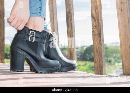 gros plan sur les pieds d'une fille latine mettant sur des bottines en cuir synthétique noir, debout sur un balcon avec des rampes en bois Banque D'Images