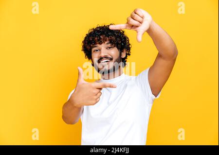 Joyeux joyeux joyeux garçon millénaire indien ou arabe en t-shirt blanc de base, faisant cadre imaginaire de l'appareil photo prise de vue, debout sur un arrière-plan orange isolé, regarde l'appareil photo, souriant Banque D'Images