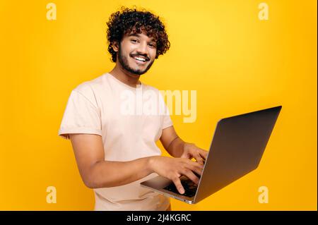 Un homme indien ou arabe millénaires aux cheveux maurires positifs dans un t-shirt, dactylographiant sur un clavier d'ordinateur portable, se tenant sur un arrière-plan orange isolé, regardant l'appareil photo, souriant amical. Concept de technologie Banque D'Images
