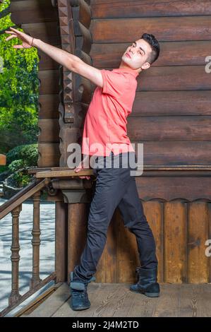 Habillée d'un polo rouge, d'un pantalon noir et de bottes en cuir, d'un bras étirant et de la rotation des jambes, un jeune homme beau est debout par un vieil homme fashio Banque D'Images