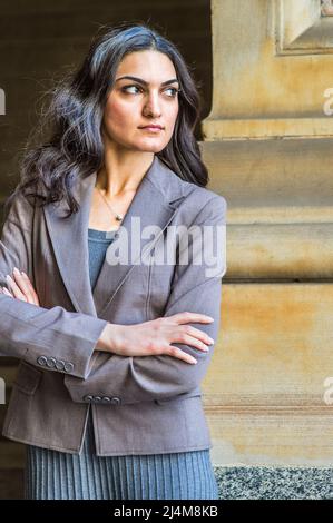 Vêtue d'une robe en tricot gris et d'une veste, croisant les bras, une jeune femme d'affaires se tient à l'extérieur, regardant avec assurance vers l'avant Banque D'Images