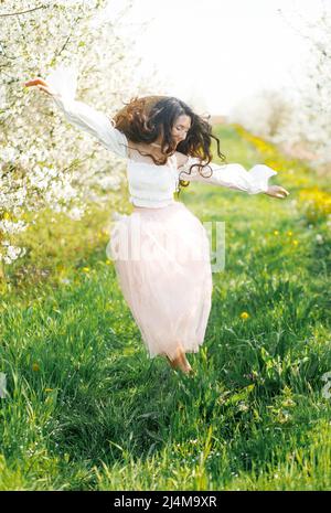 Une jolie brunette dans une jupe de tulle rose traverse un verger de cerisiers en fleurs Banque D'Images