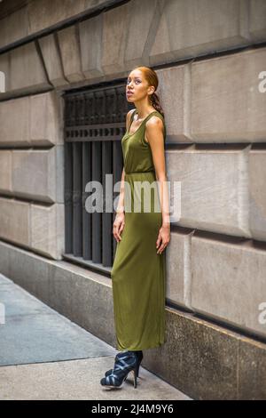 Femme se détendant dans la rue. Habillez dans un vert, longue robe maxi sans manches, noir robe sandales, une jeune femme noire est debout à côté de la vieille fenêtre de style de mode Banque D'Images