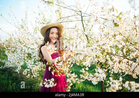 Portrait d'une fille rêveuse dans le parc de printemps parmi les arbres blancs en fleurs au coucher du soleil. Femme en chapeau de paille et robe pourpre, cerisiers en fleurs. Ressort s Banque D'Images