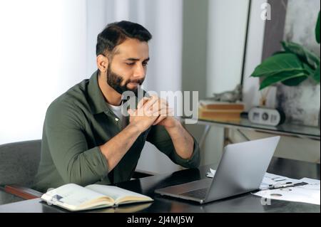 Pensive focalisé Indien ou arabe, homme à succès, courtier, gestionnaire, assis à un bureau dans le bureau, regarde attentivement un écran d'ordinateur portable, planifie une stratégie financière, évalue les risques, pense Banque D'Images