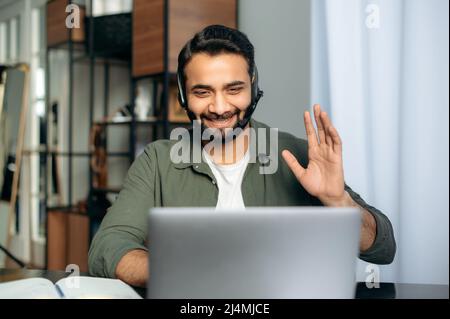 Un homme indien attrayant positif dans les écouteurs, l'opérateur de la ligne d'assistance, un employé de soutien, un consultant en ligne assis à un ordinateur portable dans le bureau, travaille avec un client sur appel vidéo à l'aide d'un casque, geste de salutation Banque D'Images