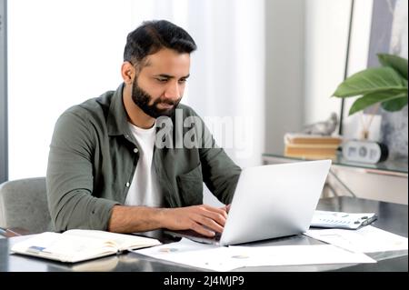 Homme indien confiant concentré utilisant un ordinateur portable dans un bureau moderne. Homme d'affaires réussi, indépendant, Manager, dans une chemise décontractée, navigation e-mail, travailler et taper sur un ordinateur portable, travailler sur un projet Banque D'Images