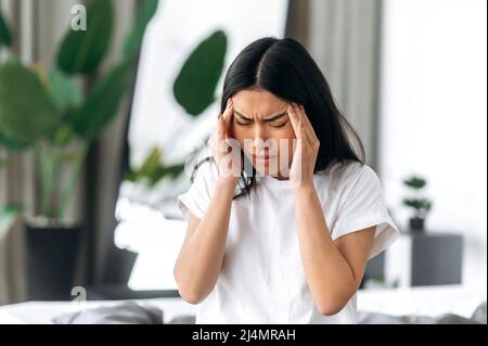 Bouleversé asiatique épuisé fille en t-shirt blanc, assis dans une chambre sur un lit, massages de ses temples, fermé ses yeux, souffre de maux de tête, migraine, travail excessif, spasme de la tête, a besoin de traitement, de repos et de sommeil Banque D'Images