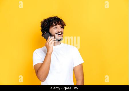 Conversation téléphonique agréable. Un homme indien ou arabe positif à poil dur dans un t-shirt, parle sur le smartphone avec un ami ou une famille, souriant, debout sur un arrière-plan orange isolé Banque D'Images