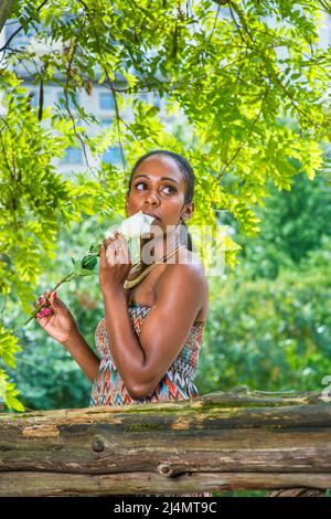 Habillant dans une robe sans bretelles colorée, portant un anneau de cou doré, une jolie femme noire porte une fleur blanche touchant sa bouche, pensant, looki Banque D'Images