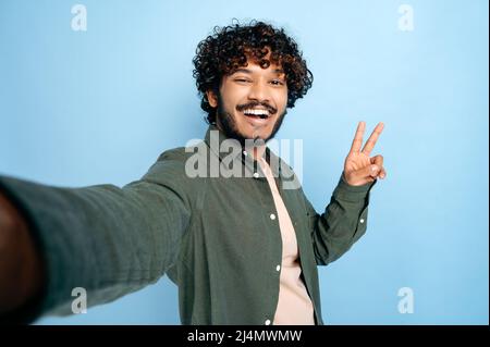 Joyeux indien ou arabe gars excités avec l'expression du visage gladen, dans des vêtements décontractés, faisant selfie tourné sur smartphone et sourires dans l'appareil photo, montre le symbole de paix, isolé fond bleu Banque D'Images