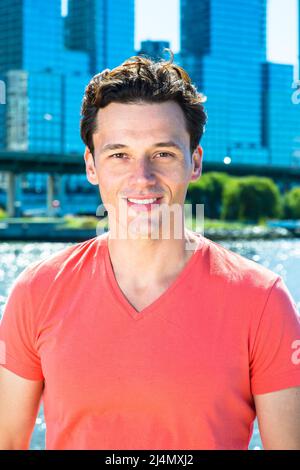 Avec un T-shirt rouge clair, un jeune homme beau est debout au bord de la rivière, vous regardant avec un sourire. Banque D'Images