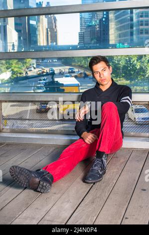 Jeune homme portant un pull à col châle noir, un Jean rouge, des chaussures en cuir noir, assis au sol près d'un grand mur de verre à New York, reposant, pensant. Banque D'Images