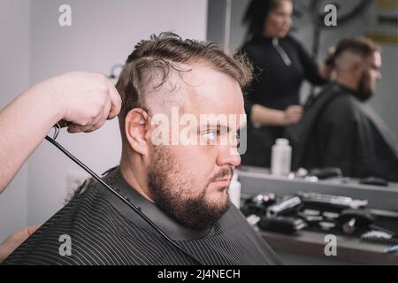 Un jeune barbu avec un mohawk se coupe la tête par un coiffeur assis dans une chaise au salon de coiffure. Rasez votre tête chauve Banque D'Images