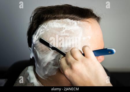Jeune homme plein de confiance d'apparence brutale du Caucase, qui se déchue de sa barbe et de ses cheveux sur la tête. Mousse de rasage à lame de rasoir dangereuse Banque D'Images