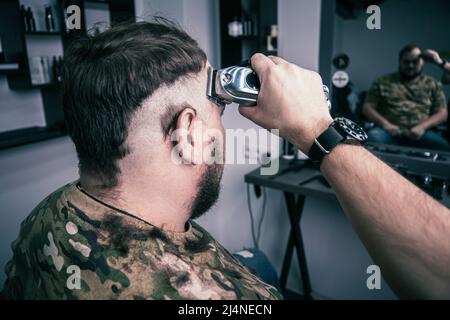 Un jeune homme dans un uniforme militaire se prépare à la tête chauve pour le service militaire. Un homme avec une barbe obtient une coupe de cheveux dans un salon de coiffure. Banque D'Images