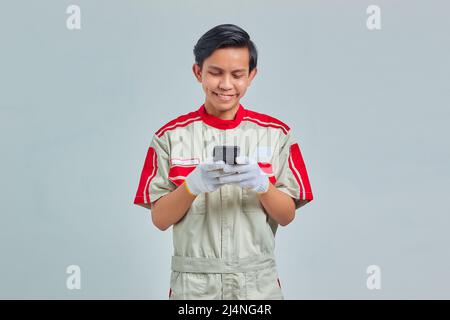 Homme souriant et beau portant un uniforme mécanique à l'aide d'un téléphone cellulaire isolé sur fond gris Banque D'Images