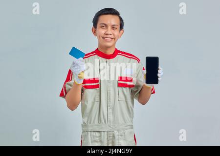 Portrait d'un jeune mécanicien souriant montrant une carte de crédit et tenant le smartphone sur fond gris Banque D'Images