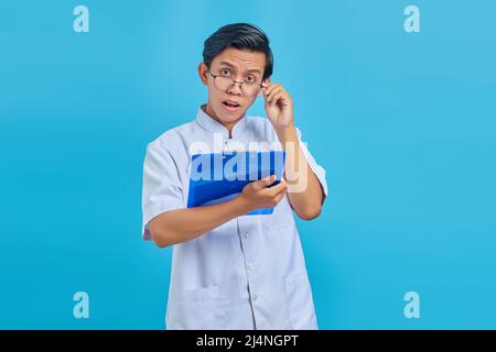 Surpris jeune infirmier asiatique prenant des lunettes et regardant des notes sur le presse-papiers avec la bouche ouverte sur fond bleu Banque D'Images