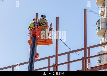 Les constructeurs masculins travaillent en hauteur dans un berceau de levage, créant le cadre en fer du bâtiment. Banque D'Images