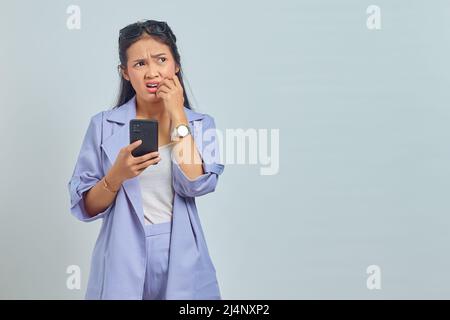 Portrait d'une jeune femme asiatique utilisant un téléphone cellulaire, aspect stressé et nerveux avec la main dans la bouche mordant l'ongle sur fond blanc Banque D'Images