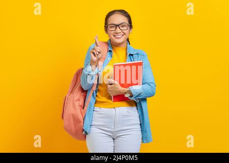 Portrait de la jeune femme asiatique heureuse étudiant en vêtements denim avec sac à dos tenant carnet, optimiste et plein d'espoir, croiser les doigts pour bonne chance isola Banque D'Images