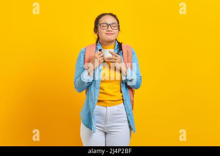 Portrait d'une jeune femme asiatique en vêtements denim avec sac à dos tout en appréciant une tasse de café isolée sur fond jaune Banque D'Images