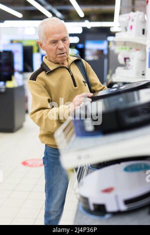 un homme âgé aux cheveux grisés qui regarde le robot hoover au comptoir dans la salle d'exposition du département d'hypermarché des appareils électriques Banque D'Images