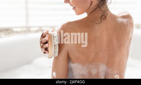 Vue arrière de la femme millénaire faisant un massage lymphatique avec la brosse, frottant la peau tout en prenant le bain de bulles à la maison Banque D'Images