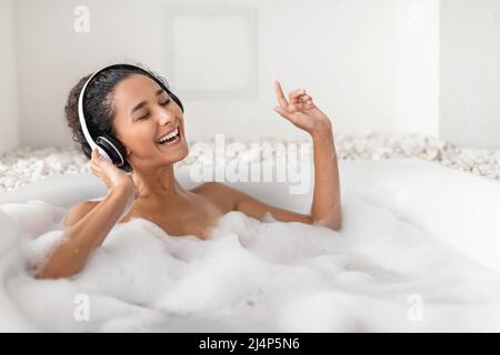 Une jeune femme insouciante dans un casque qui écoute de la musique, chante votre chanson préférée tout en étant allongé dans un bain bouillant à la maison Banque D'Images