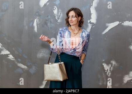 Fille tendance et élégante posant dans des pontons verts, chemisier multicolore contre un mur gris Banque D'Images