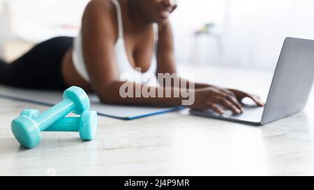 Une jeune femme noire plousse, allongé sur un tapis de yoga et regardant des vidéos de sport en ligne sur un ordinateur portable à la maison, se concentre sur les haltères Banque D'Images