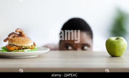 Femme noire en surpoids regardant le hamburger sous la table, en choisissant entre la nourriture saine et malsaine à la maison Banque D'Images