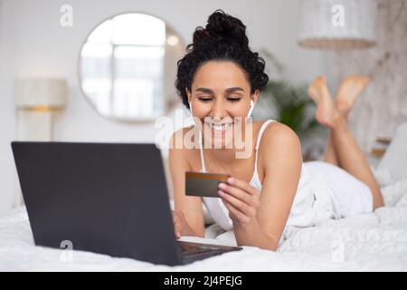 Satisfaite jeune caucasien brune femelle avec cheveux bouclés en pyjama payant de l'argent avec carte de crédit et ordinateur portable Banque D'Images