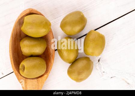 Quelques olives vertes bio piquantes et savoureuses et épicées et une cuillère en bois, sur fond de bois peint blanc naturel, vue du dessus. Banque D'Images