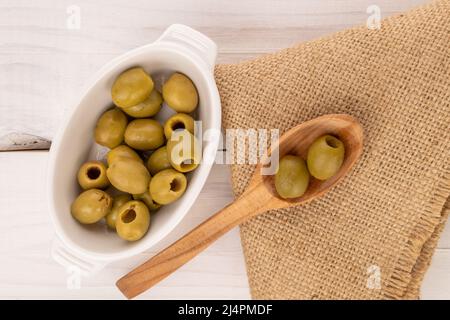 Quelques olives vertes piquantes bio savoureuses et épicées se trouvent sur une soucoupe en céramique blanche, une cuillère en bois et une serviette en jute sur fond de peinture blanche naturelle Banque D'Images