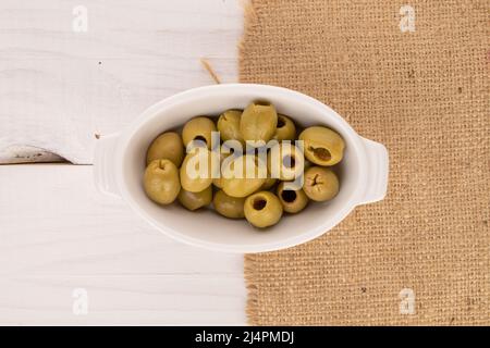 Quelques olives vertes piquantes bio épicées se trouvent sur une soucoupe en céramique blanche, une serviette en jute sur un fond de bois peint blanc naturel, vue du dessus Banque D'Images