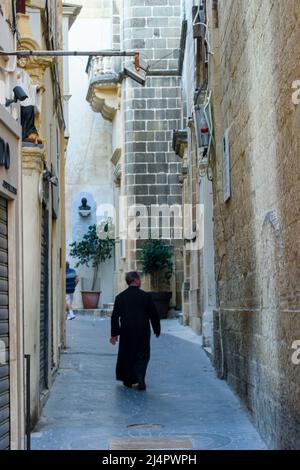 Un prêtre portant une cassock noire s'émène dans une rue étroite à Victoria, Gozo, Malte. Banque D'Images