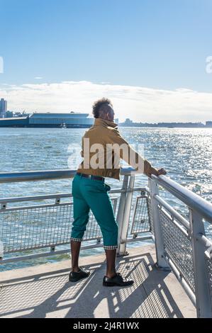 Portant une veste en laine jaune foncé, un pantalon vert et des chaussures en cuir noir, un jeune homme noir aux cheveux mohawk est debout près d'une rivière et regarde Banque D'Images