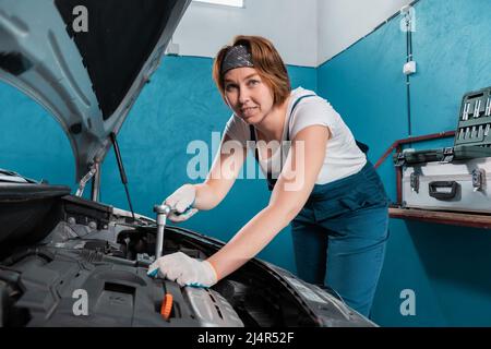 Femme de mécanicien souriante en combinaison bleue réparer un moteur avec clé à cliquet. Le concept de l'égalité de la femme et travaille à l'atelier de réparation automobile. Banque D'Images