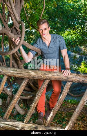 Portant une chemise Henley à manches longues grises, un Jean rouge et des chaussures marron, un jeune homme se tient derrière une clôture en bois, un angle de bras Banque D'Images