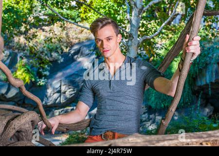 Portant une chemise Henley à manches longues grises et patte roulée, un jeune homme charmant est debout dans une zone de bois, tenant des hotons, avec charme vous regarder. Banque D'Images