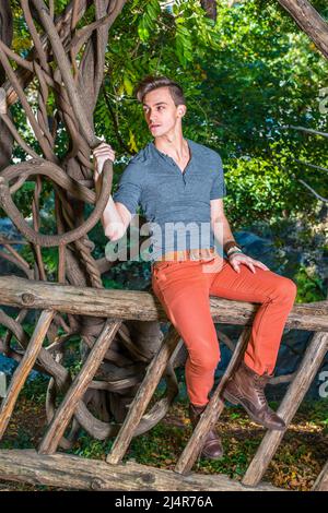 Portant une robe dans une manche longue grise avec une chemise Henley à patte rouillée, un Jean rouge, des chaussures de bottes en cuir marron, un jeune homme est assis sur une clôture en bois, tenant un Banque D'Images