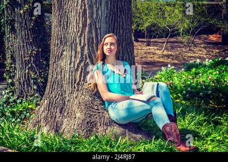 Adolescente américaine portant un haut sans manches bleu, un Jean tendance, des bottes brunes, assis contre le tronc d'arbre à Central Park, New York, lisant rouge b